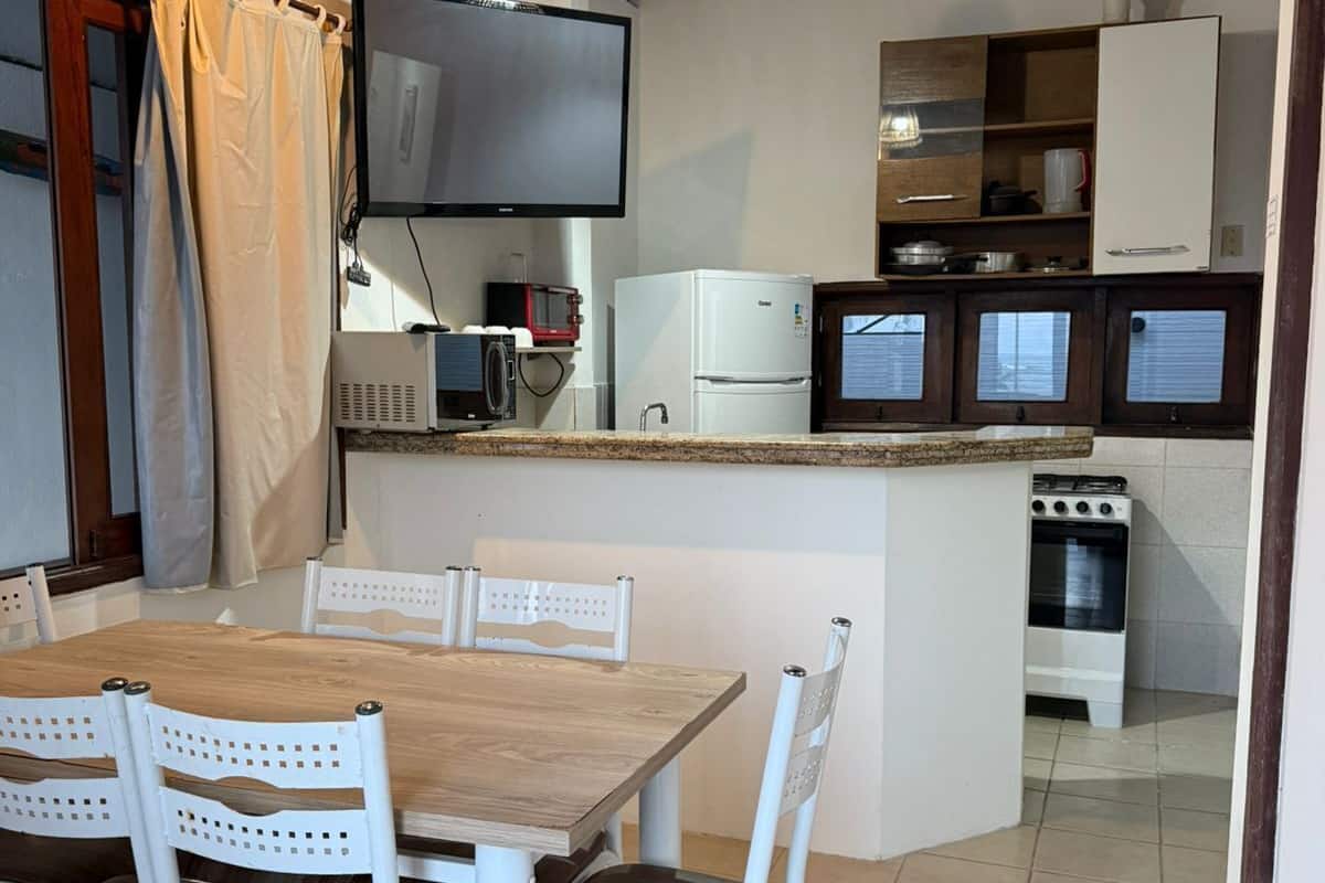 A compact kitchen with a dining area, including a wooden table, white chairs, a TV, a microwave, a refrigerator, and a small oven.