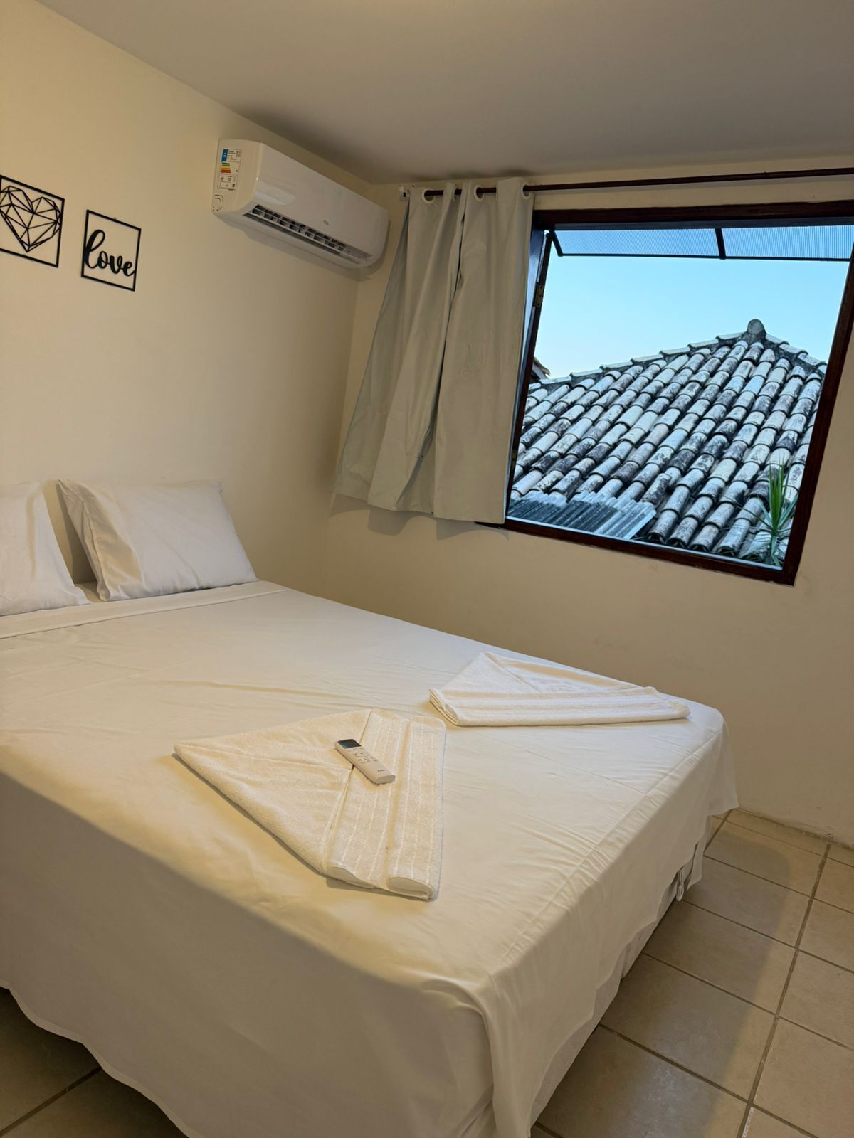 Uma cama bem arrumada com controle remoto e toalhas dobradas, um ar condicionado fixado na parede e uma janela com vista para um telhado em cerâmica.