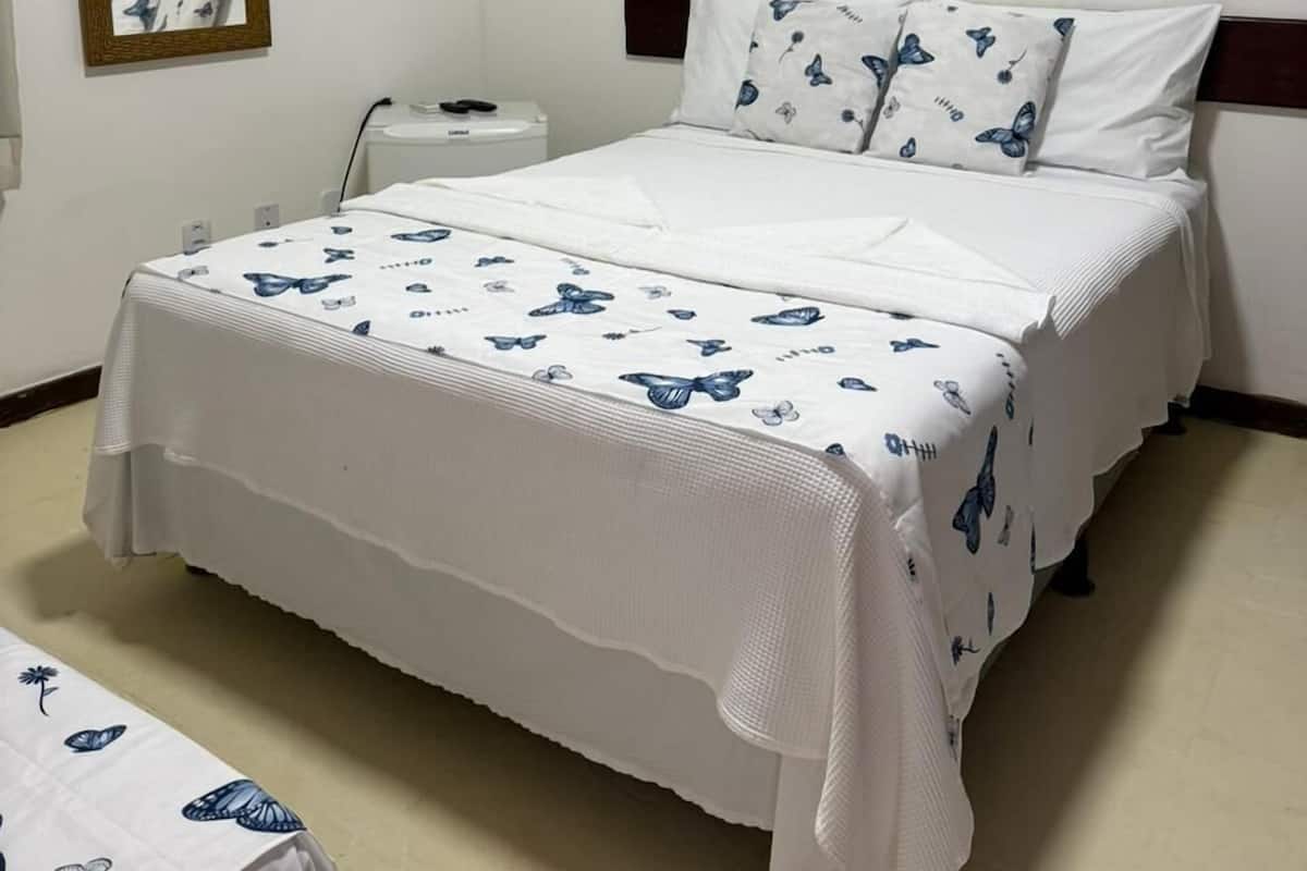 A hotel room with a bed, a bedside table, an air conditioner, a mirror, and a painting on the wall.