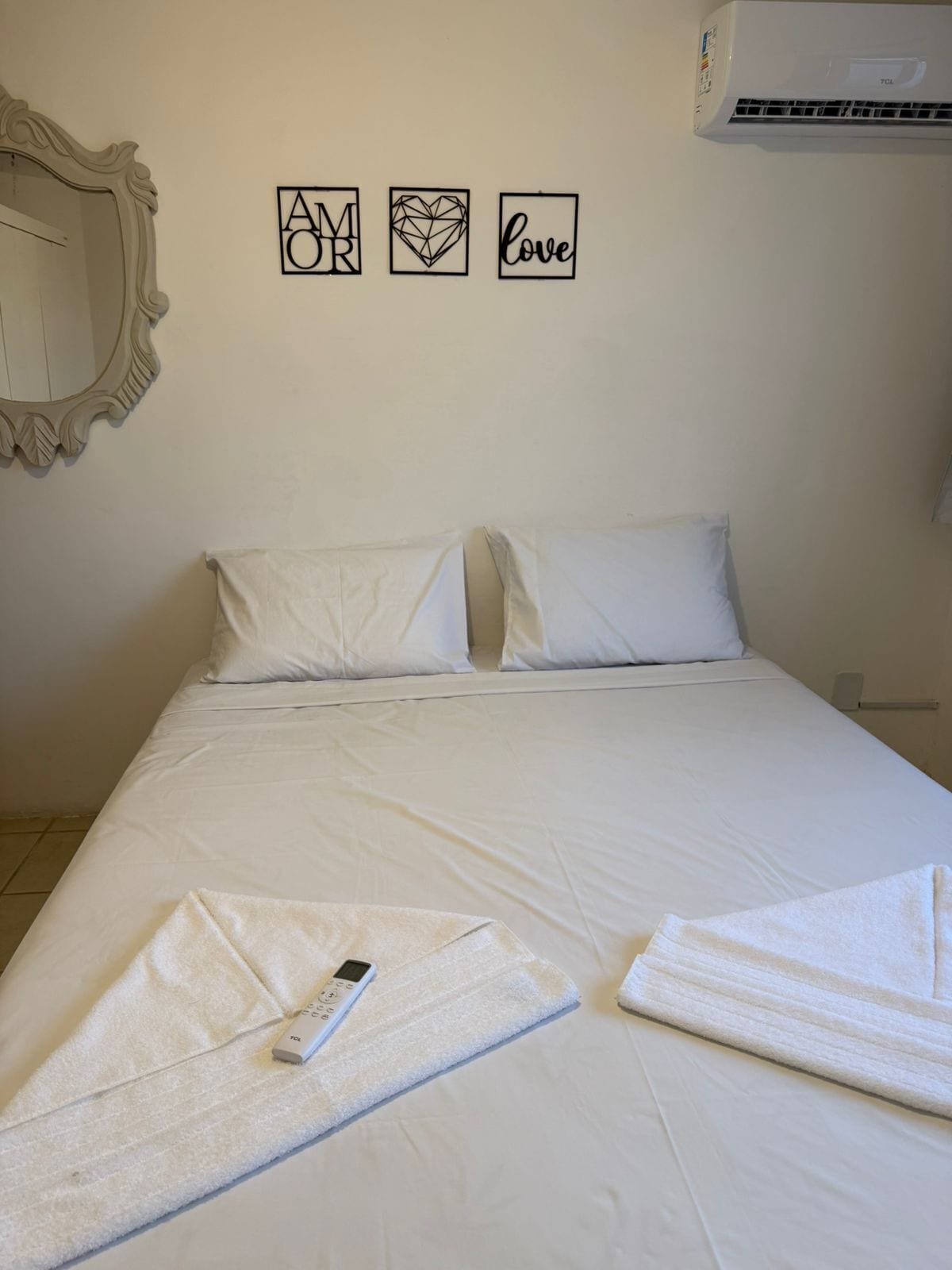 A neatly made bed with white linens, a folded towel, and a remote control on the bed. There are three wall decorations above the bed: a square with the letters "AM OR", a heart-shaped decoration, and the word "love".