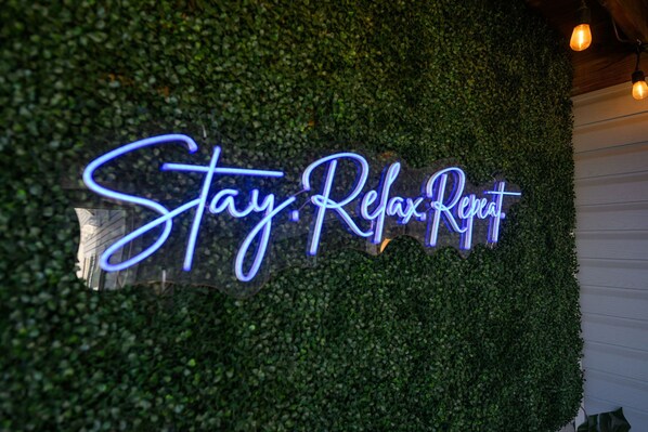 A neon sign with the text 'Stay. Relax. Rent.' against a backdrop of green foliage.