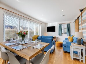 A modern living room with a wooden dining table, blue sofas, and a TV mounted on the wall.