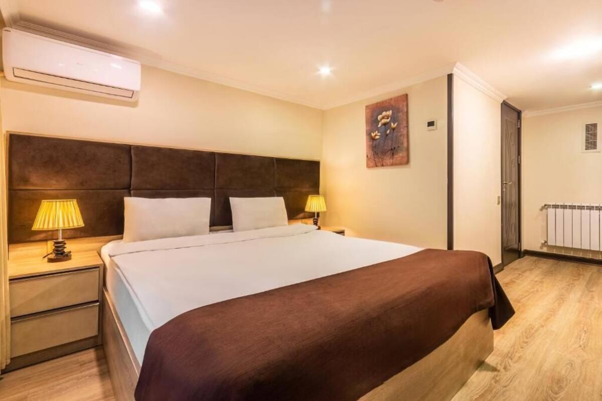 A hotel room with a large bed, two bedside lamps, a wall-mounted air conditioner, and a radiator.