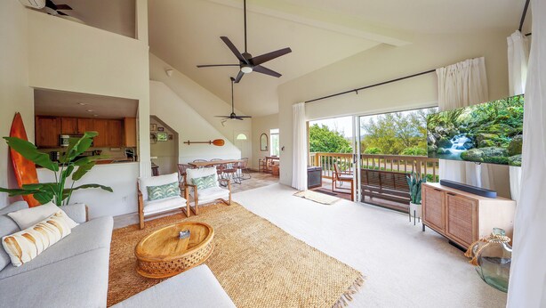 A spacious living area with a large sofa, a coffee table, and a dining area with chairs. There is a balcony with a view of greenery.
