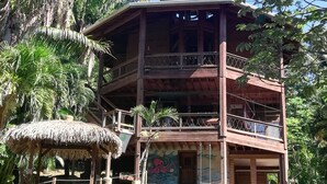 A multi-level wooden building with balconies, surrounded by lush greenery and a thatched structure.
