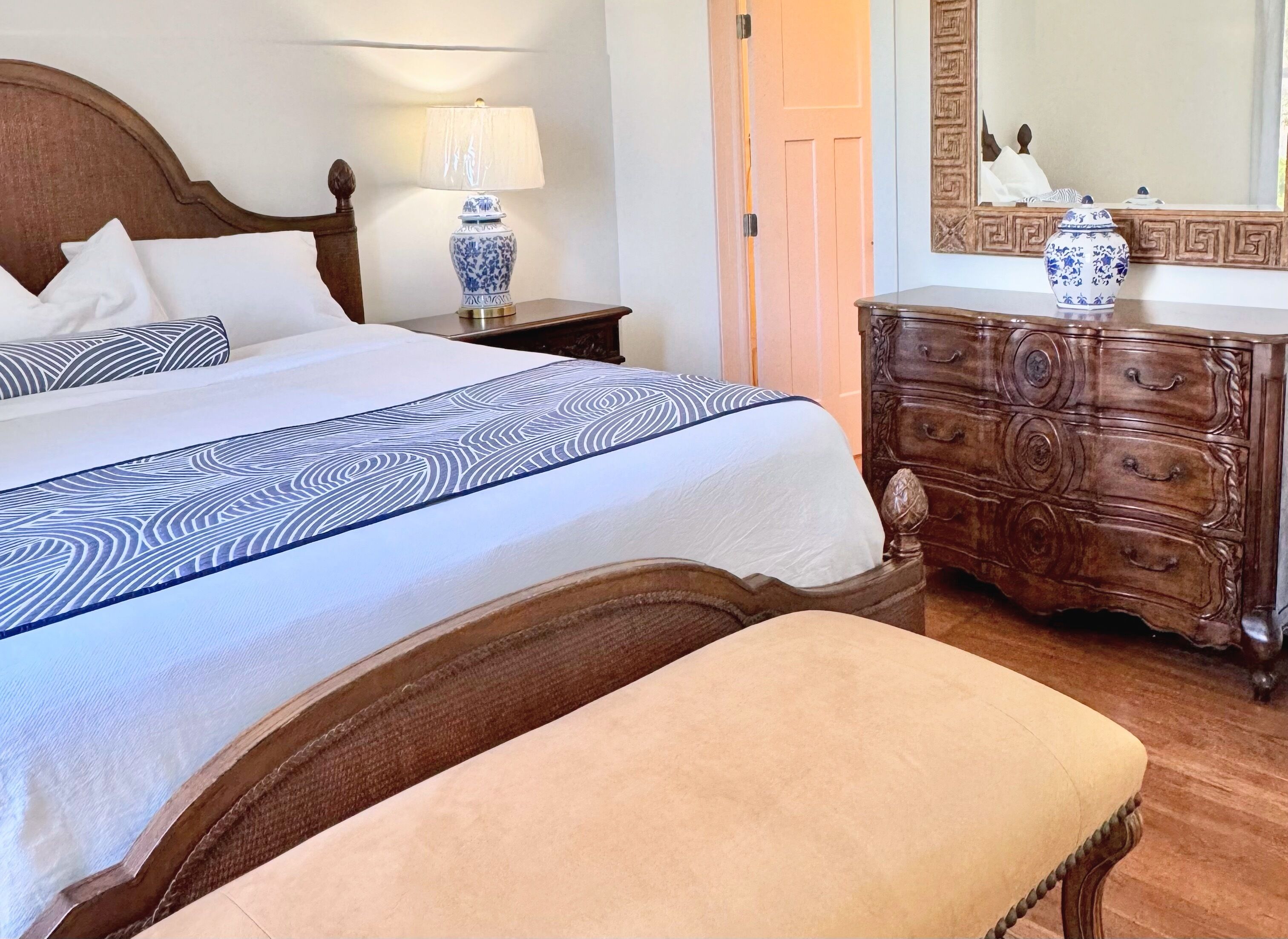 A neatly made bed with a patterned bedspread, a bedside table with a lamp, a wooden dresser, and a bench.