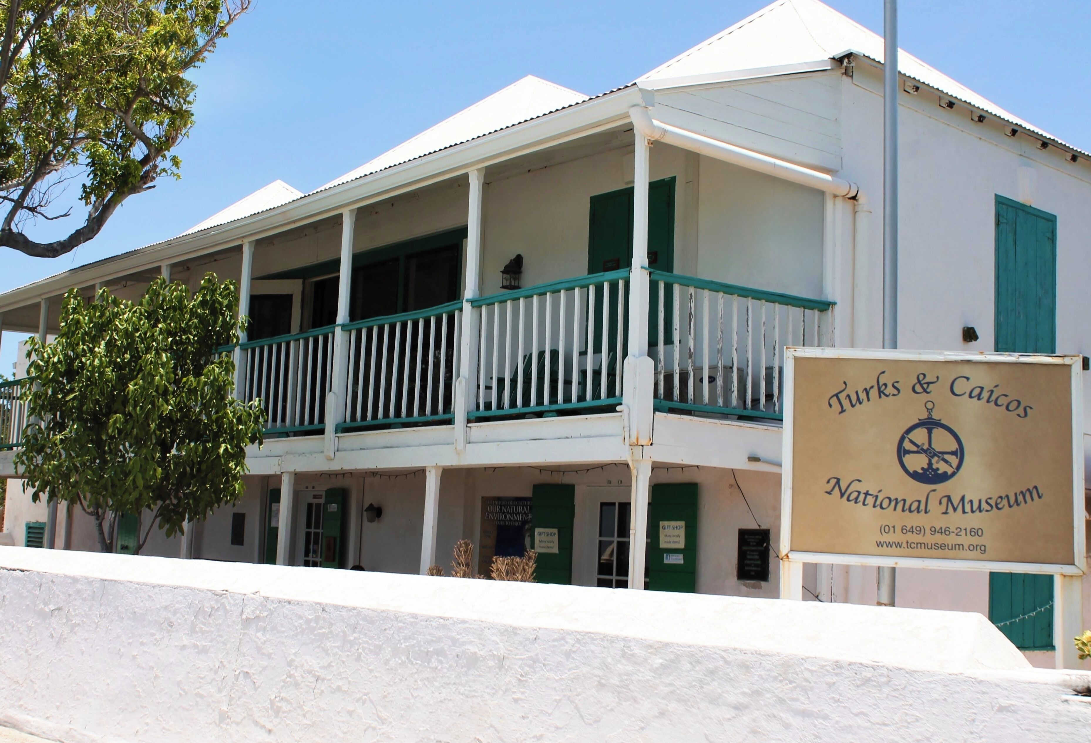 A two-story building with a balcony, a sign for the Turks & Caicos National Museum, and a tree in front.
