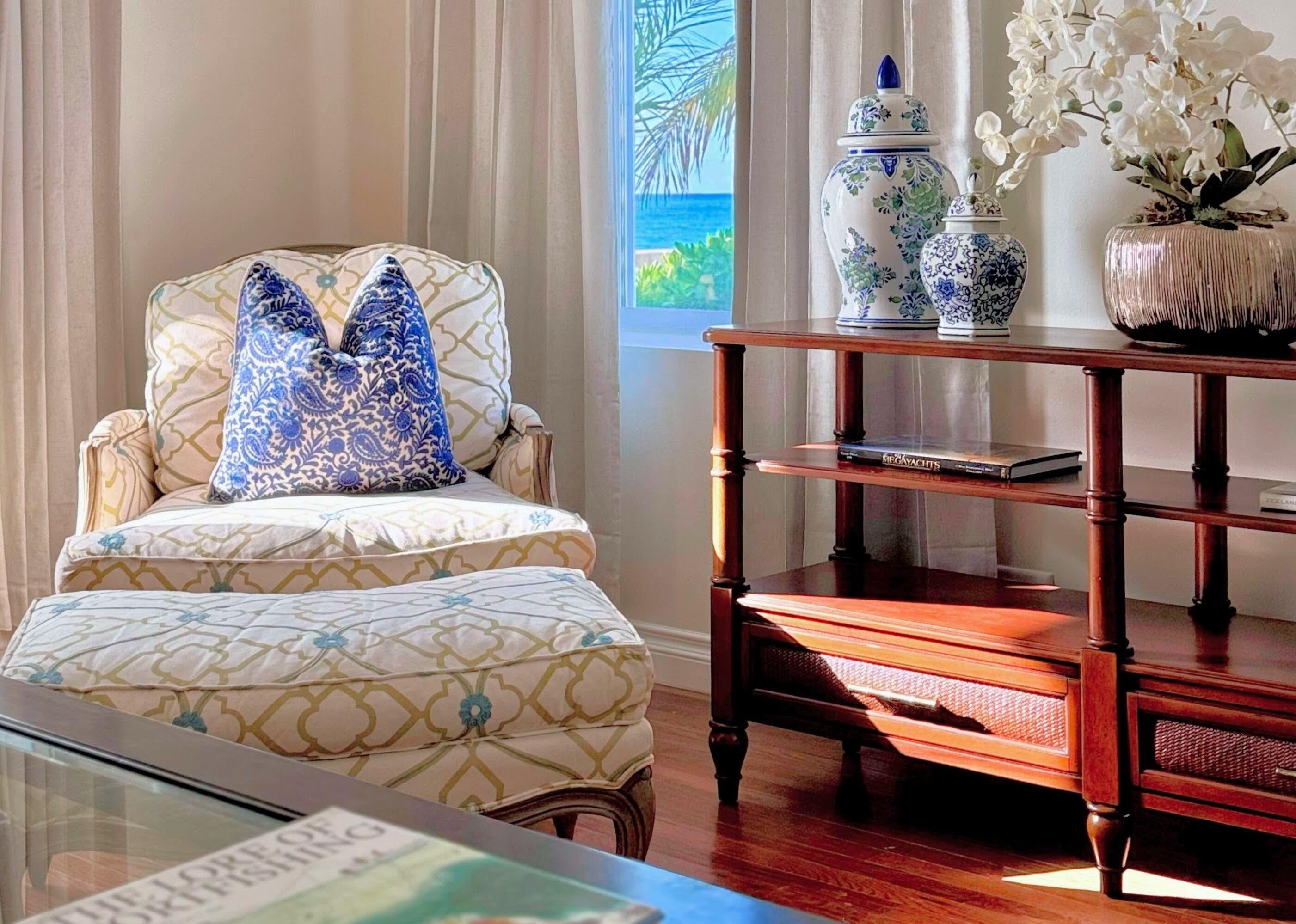 A room with a patterned armchair, a wooden side table with books, and a view of the ocean through a window.