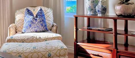 A room with a patterned armchair, a wooden side table with books, and a view of the ocean through a window.