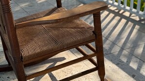 Ein Holzhocker mit gewebter Sitzfläche auf einem Balkon mit weißer Brüstung, mit Blick auf ein Gewässer und viel Grün.
