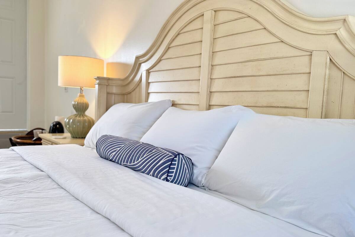 A neatly made bed with white linens and a striped pillow, a wooden headboard, and a bedside lamp on a nightstand.