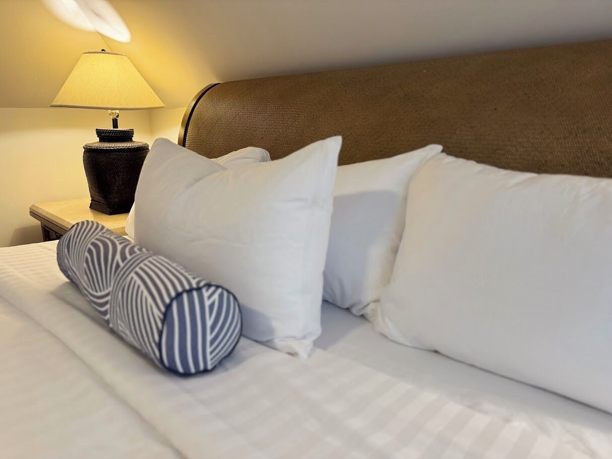 A neatly made bed with white bedding and striped pillows, a brown headboard, and a bedside lamp.