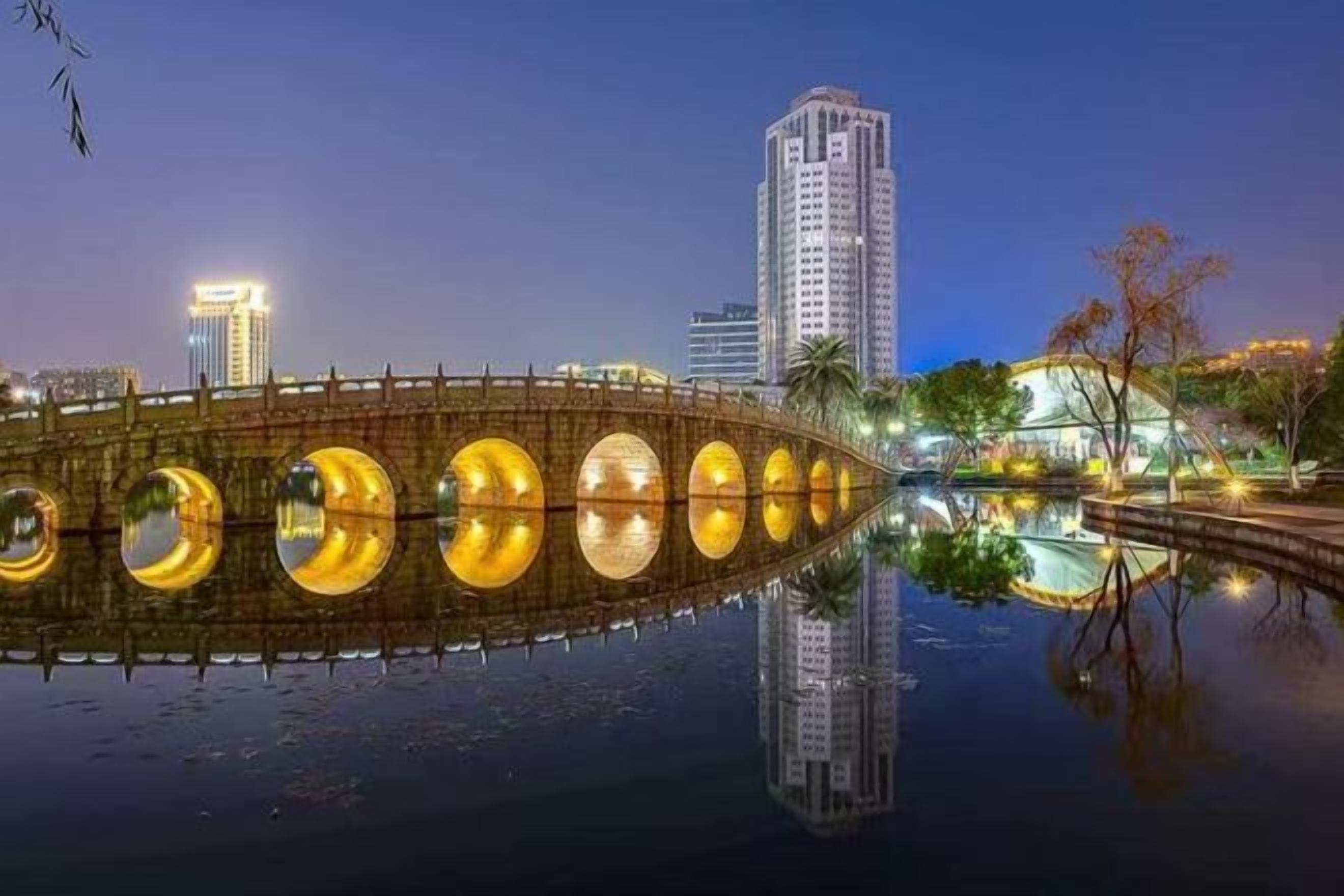 A bridge with circular lights reflecting on the water, surrounded by modern buildings.