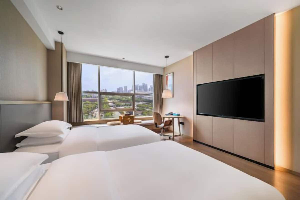 A hotel room with two beds, a desk, a TV, and a large window offering a city view.