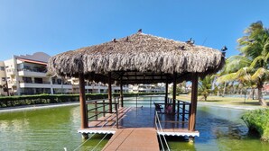 Een houten brug leidt naar een paviljoen met rieten dak boven een waterpartij, omringd door groen en gebouwen.