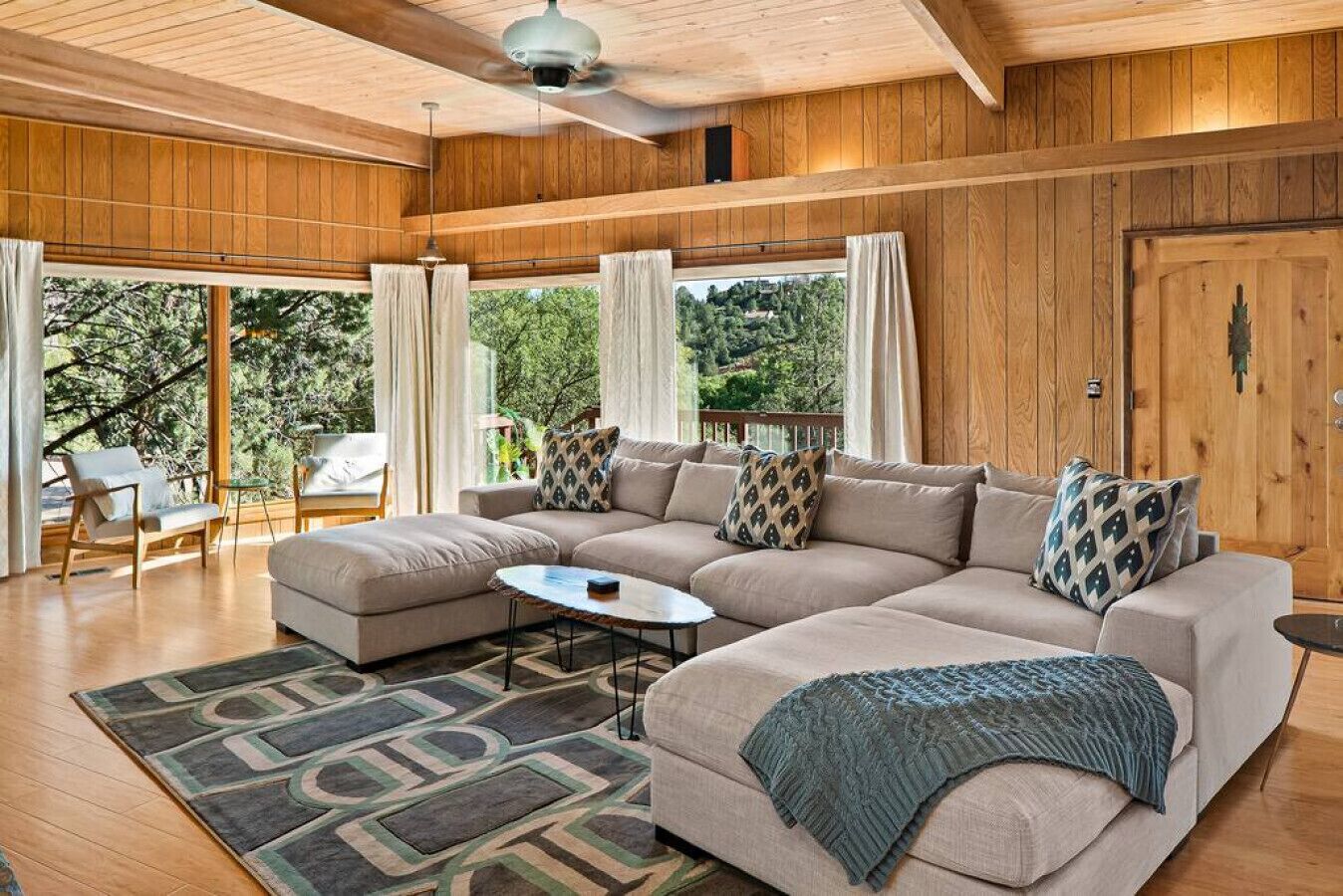 A spacious living room with a large L-shaped sofa, a coffee table, and a patterned rug. There are two chairs and a small table with a lamp. The room has wooden walls and a large window with curtains.