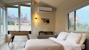 A bedroom with a bed, a bench, a wall-mounted air conditioner, and a large window with a view of red rock formations.