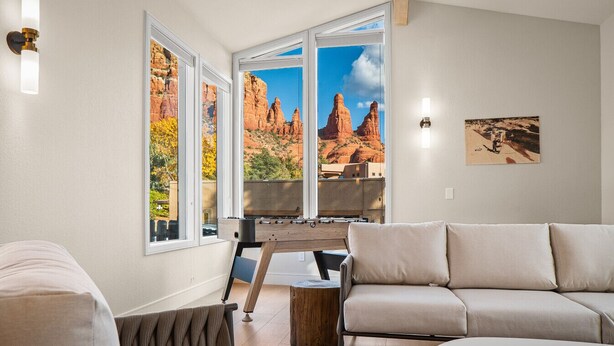 A living room with a sofa, a coffee table, and a foosball table. There are large windows with a view of red rock formations.