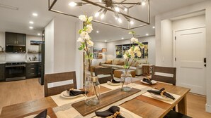 A modern dining area with a wooden table set for four, surrounded by chairs. There is a kitchen in the background with dark cabinets and stainless steel appliances.