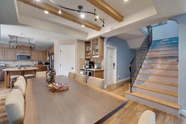 A spacious kitchen with wooden beams, a dining area with a large table, and a staircase with a painted railing.