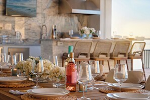Una mesa de comedor dispuesta con platos, vasos y una botella de vino, con vista al mar.
