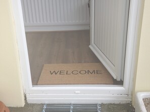 Una porta con un tappetino con la scritta 'WELCOME' e vista diretta su una stanza con pavimento in legno.