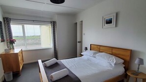 A neatly made bed with white linens, a wooden headboard, and two rolled towels. There is a small bedside table with a lamp, a wooden nightstand, and a picture on the wall. A window with curtains and a view of the outdoors is visible. - The Cottage at The Burren Tyrella - your coastal base for the Mourne country. (Downpatrick)