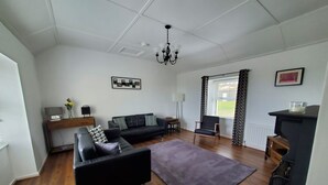 A modern living room with a black sofa, a coffee table, and a fireplace. There is a chandelier hanging from the ceiling and a window with curtains. - The Cottage at The Burren Tyrella - your coastal base for the Mourne country. (Downpatrick)