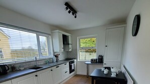 A kitchen with white cabinets, a black countertop, and a tiled floor. There is a window with blinds, a radiator, and a clock on the wall. - The Cottage at The Burren Tyrella - your coastal base for the Mourne country. (Downpatrick)