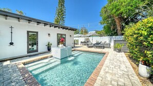 Una piscina con solárium empedrado, mobiliario de exterior y un edificio blanco con puerta y ventanas.