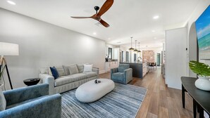 Una sala de estar moderna con un sofá, un reposapiés y sillones, una cocina al fondo y un ventilador de techo.