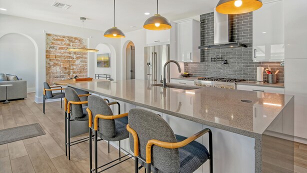 Una cocina moderna con una isla central, taburetes de bar, iluminación colgante y una pared de ladrillo decorativa.