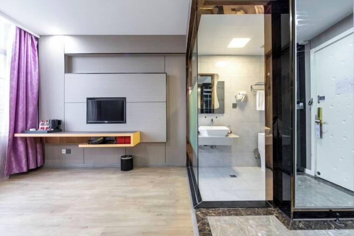 A modern hotel room with a flat-screen TV mounted on a wall, a small shelf with items, a glass shower enclosure, and a white door.