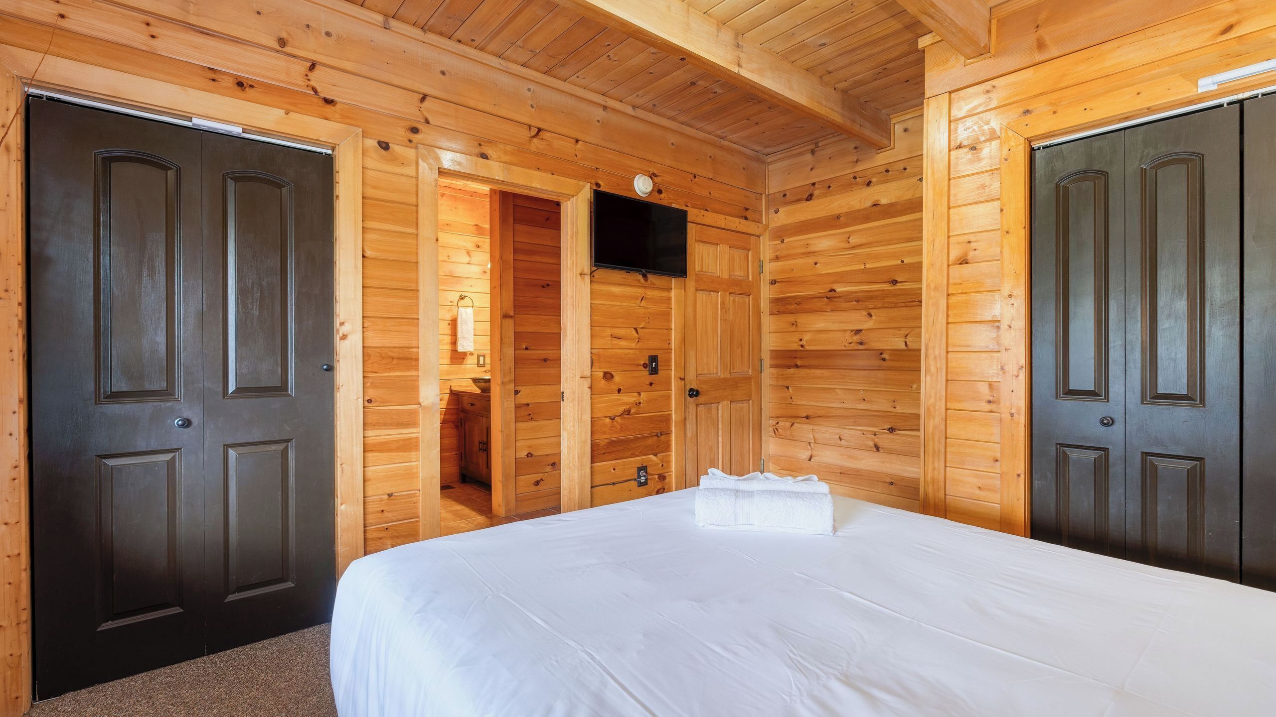 Una habitación de cabaña de madera con cama, dos puertas, un baño pequeño y un televisor.