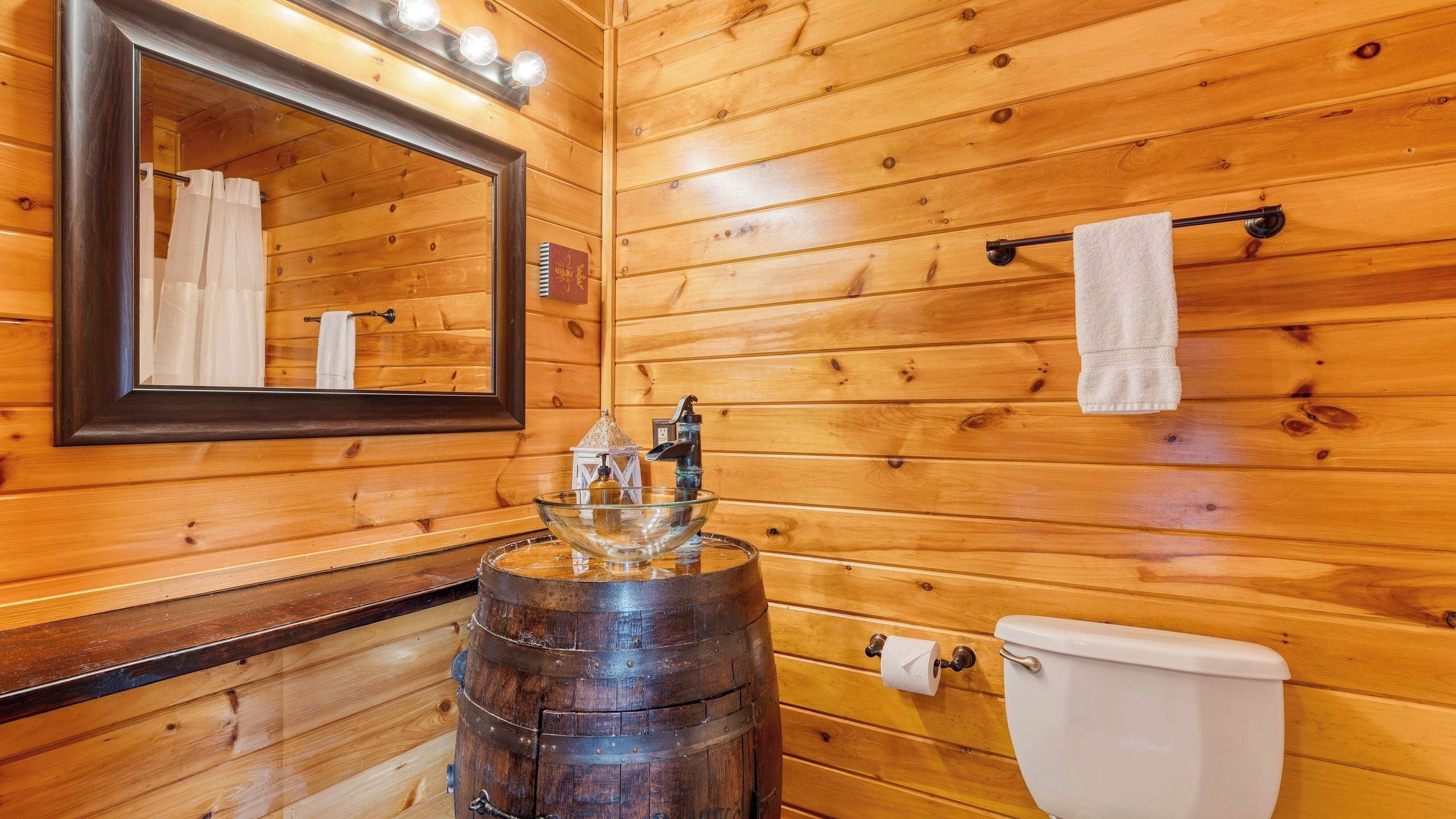 Een houten badkamer met een vakantiehuisje, een grote spiegel, een glazen wasbak, een toilet en een handdoekhouder.