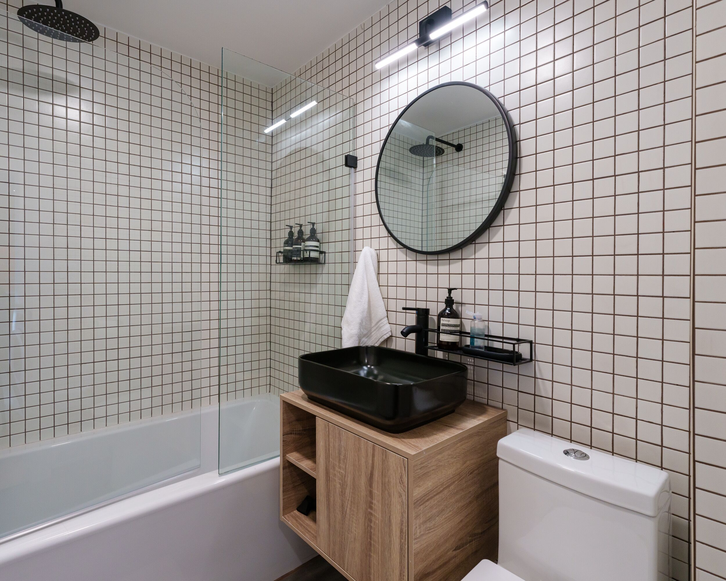 Un baño moderno con bañera, inodoro, lavamanos de cerámica negra, tocador de madera, espejo redondo y ducha.