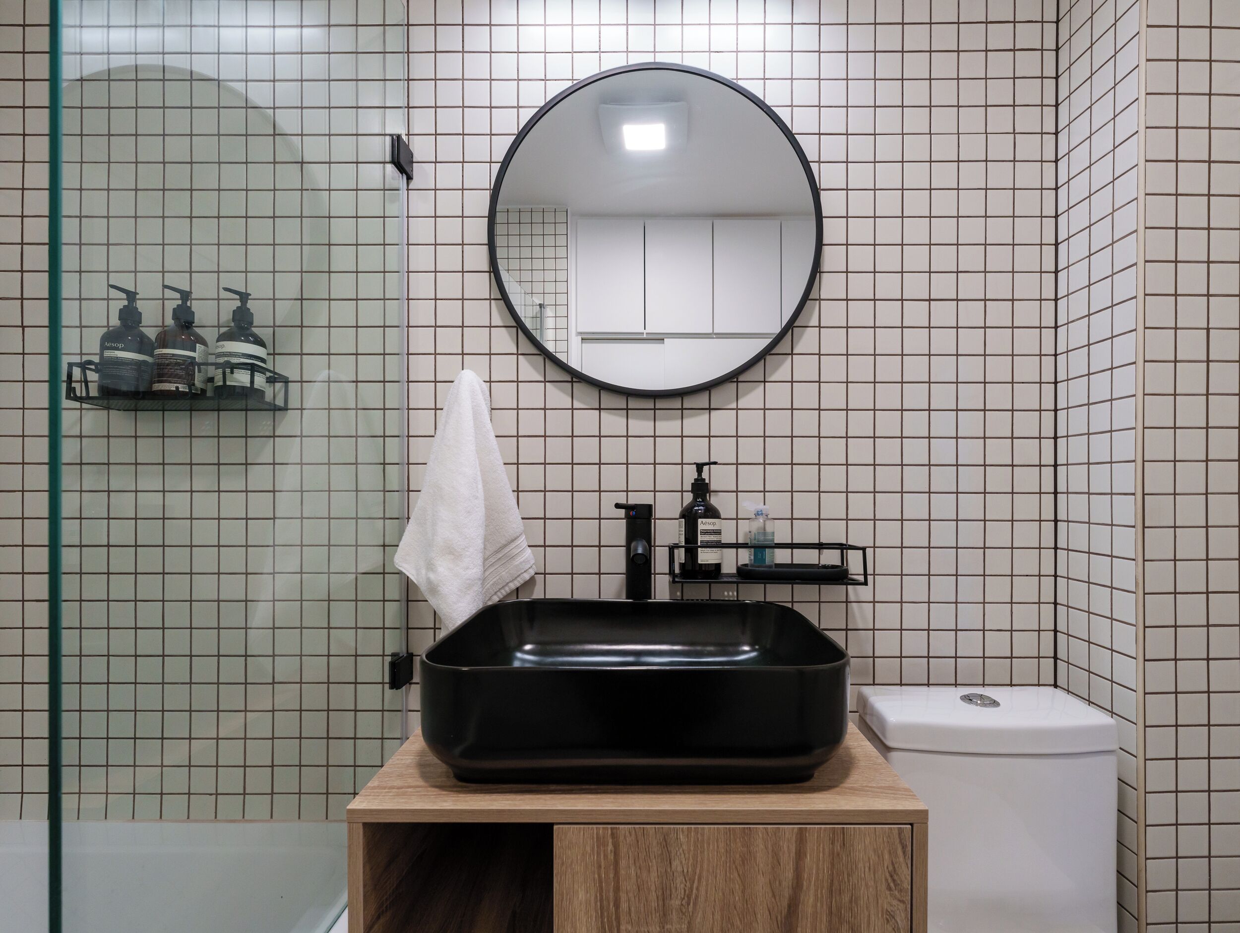 A bathroom with a black sink, a round mirror, and a white toilet. There is a towel hanging on the wall and a shelf with toiletries.