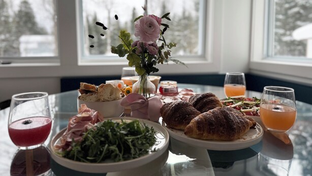 Una mesa de comedor con comida y bebidas, un florero con flores y ventanas que dan a una vista nevada.