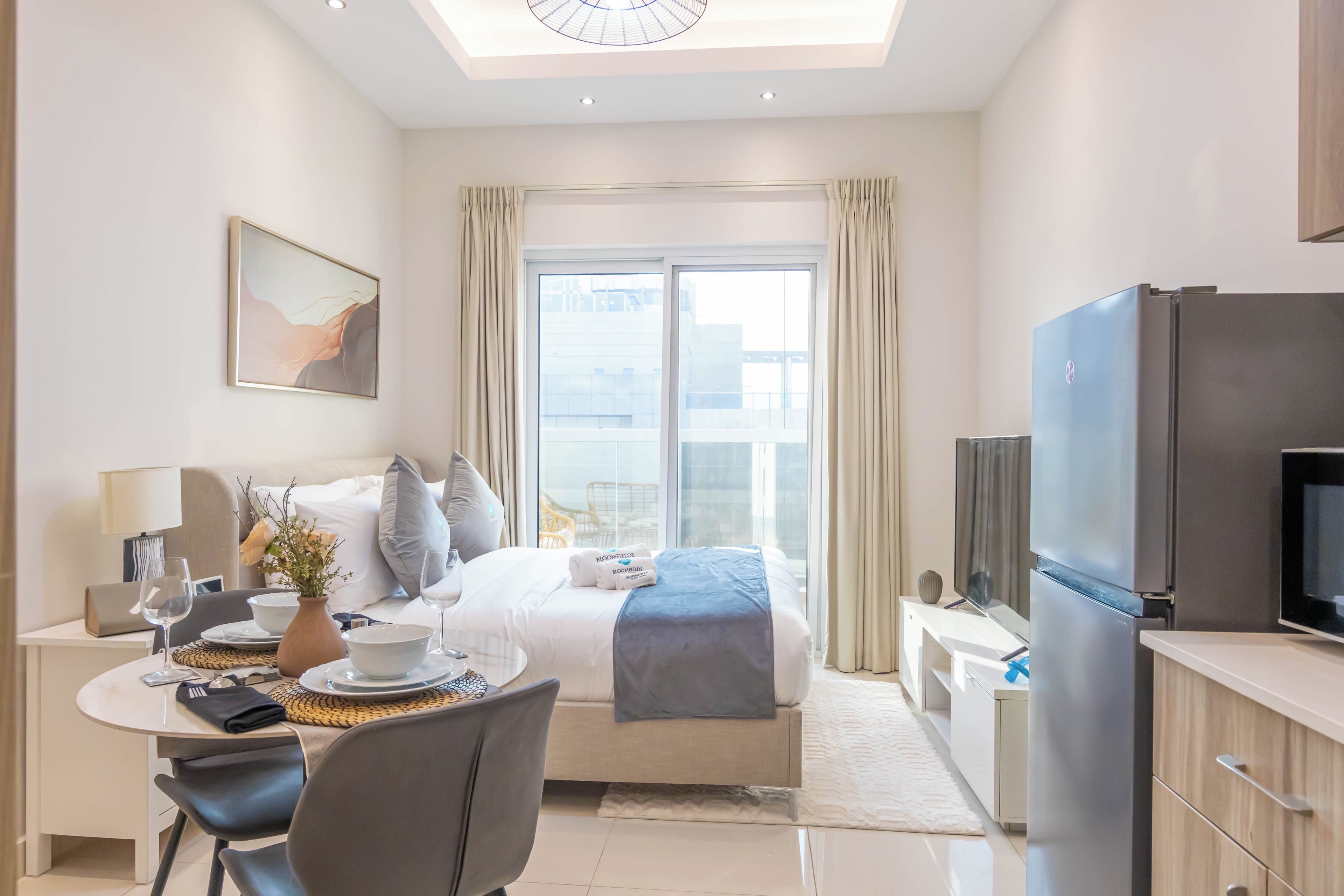 A modern hotel room with a bed, a desk with a chair, a dining table, a television, and a large window with curtains.