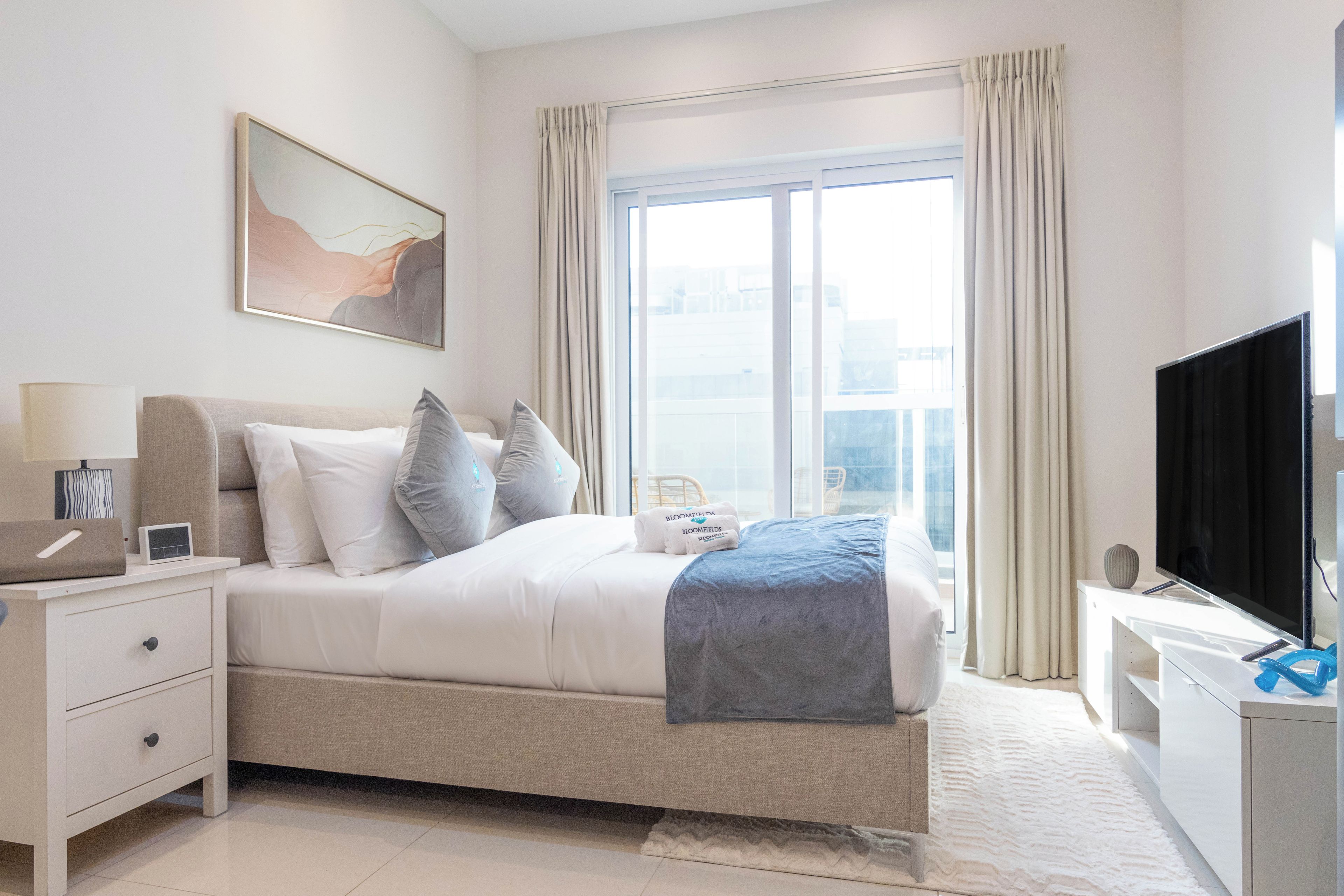 Quarto de hotel moderno com cama, mesa de cabeceira, televisão e uma janela grande com cortinas.