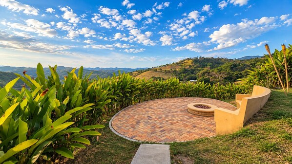 Un espacio de estar al aire libre con forma circular, piso estampado, rodeado de vegetación y vistas a colinas distantes.