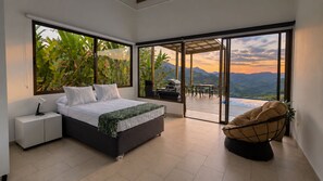 Un dormitorio con cama, mesitas de noche, una silla y vistas a montañas a través de amplias ventanas.