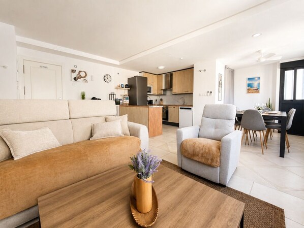 A modern living room with a sofa, armchair, and coffee table. A kitchen is visible in the background with cabinets and appliances.