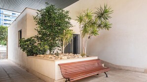 Un banc situé dans un espace extérieur abrité, doté d’un sol carrelé, et entouré de plantes ainsi que d’un petit jardin de rocaille.
