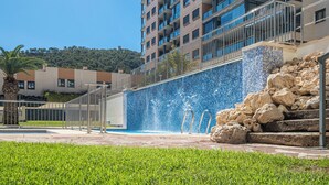 A modern building with a blue mosaic wall, a pool with a water feature, and a staircase leading to an upper level.