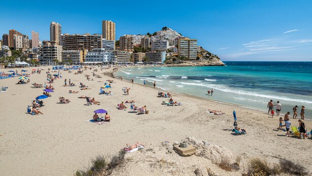 A beach with people sunbathing and swimming, a cityscape in the background, and a clear blue sky.