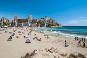 A beach with people sunbathing and swimming, a cityscape in the background, and a clear blue sky.