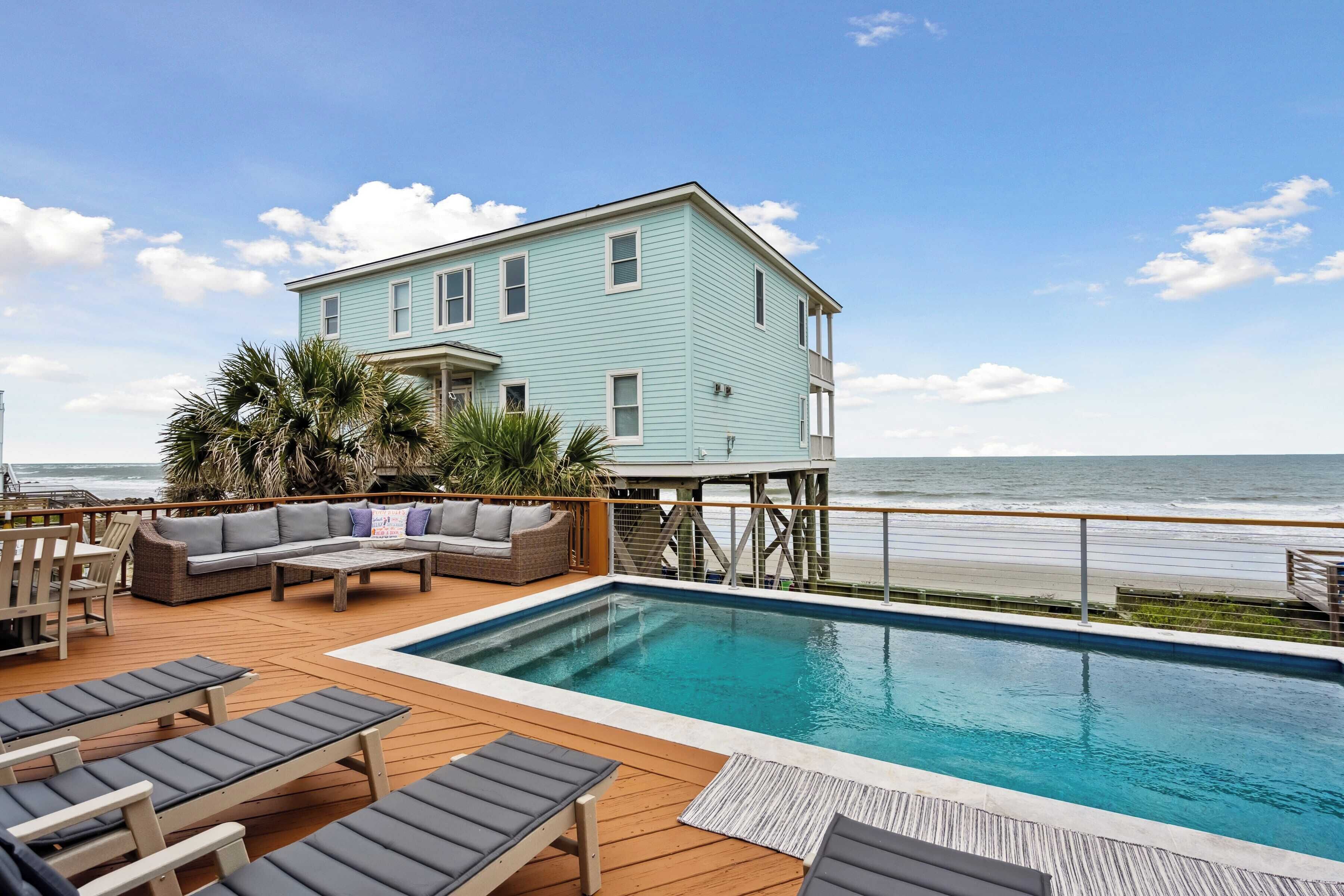 A pool area with lounge chairs, a wooden deck, and a light blue building with a balcony overlooking the ocean.