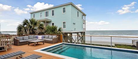 A pool area with lounge chairs, a wooden deck, and a light blue building with a balcony overlooking the ocean.