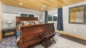 A bedroom with a wooden ceiling, a bed with plaid bedding, a nightstand with a lamp, a window with a view of mountains, and a large mirror on the wall. - Pond Property with Mountain Views (Bayfield)